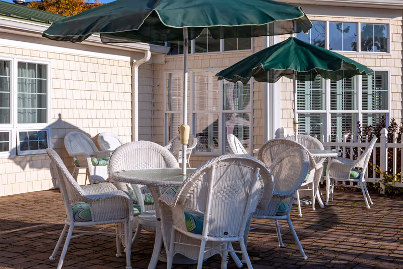 Outdoor patio with white wicker tables and chairs under green umbrellas beside a light-colored building.