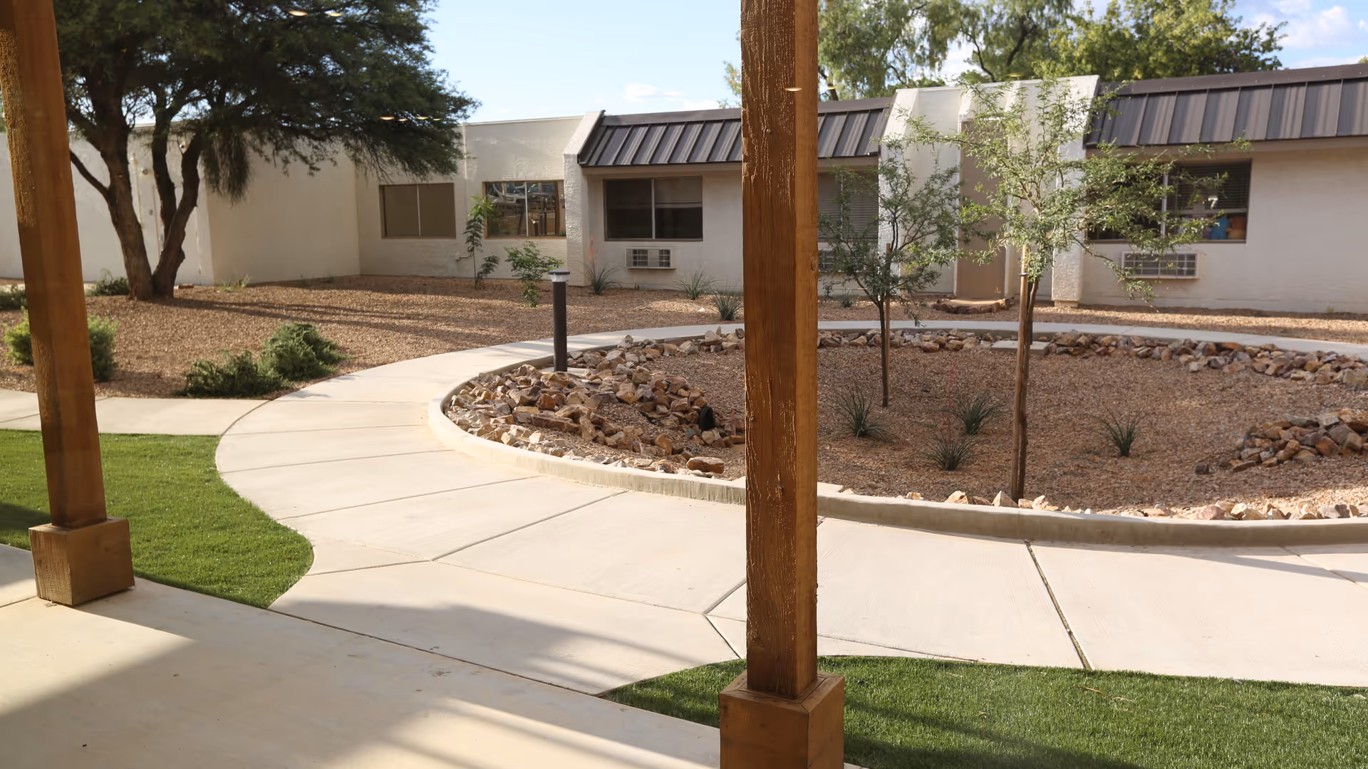Outdoor courtyard area with a curved concrete walkway, small trees, and landscaping with rocks and mulch. The courtyard is surrounded by a single-story building with windows and air conditioning units. Wooden posts support a covered patio area in the foreground.