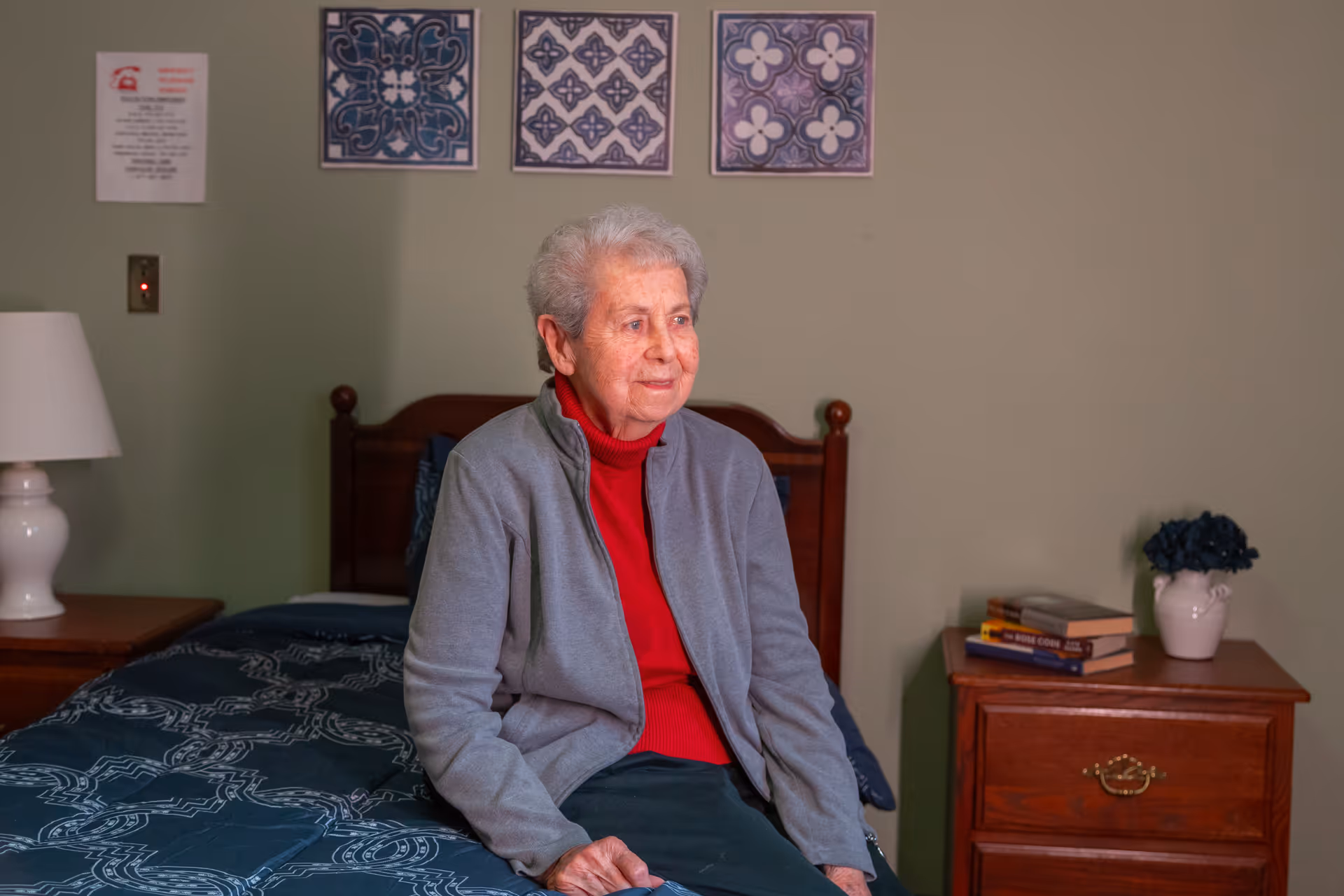 An elderly woman with short gray hair wearing a red turtleneck and gray jacket sits on the edge of a bed in a bedroom. The bed has a dark blue patterned bedspread. Behind her on the wall are three framed decorative tile artworks. To the left is a nightstand with a white lamp, and to the right is another wooden nightstand with a small vase of dark flowers and a few books stacked on it.