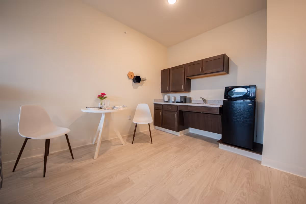 A small kitchenette area with dark wood cabinets, a black mini refrigerator, and a microwave on top. There is a white round table with two white chairs, and a small vase with pink flowers on the table. The floor is light wood, and the walls are painted a light beige color.