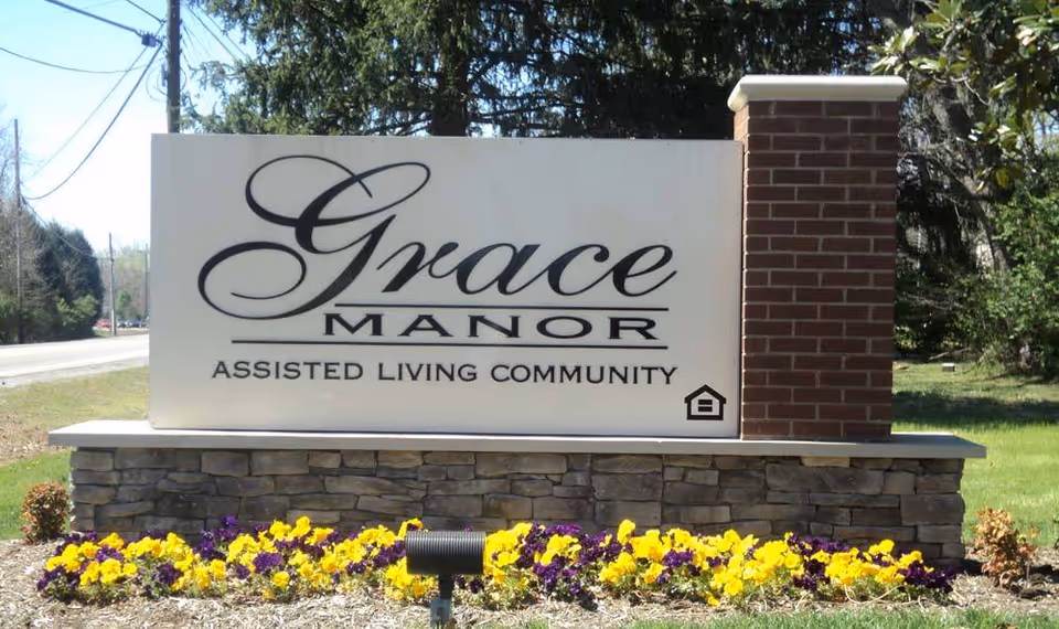 A large outdoor sign for Grace Manor Assisted Living Community, mounted on a stone and brick base with yellow and purple flowers planted in front. The sign is positioned near a road with trees and power lines visible in the background.