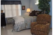 A cozy nursing facility bedroom with a single bed, bedside lamp, upholstered armchair, window with vertical blinds, and a potted plant.