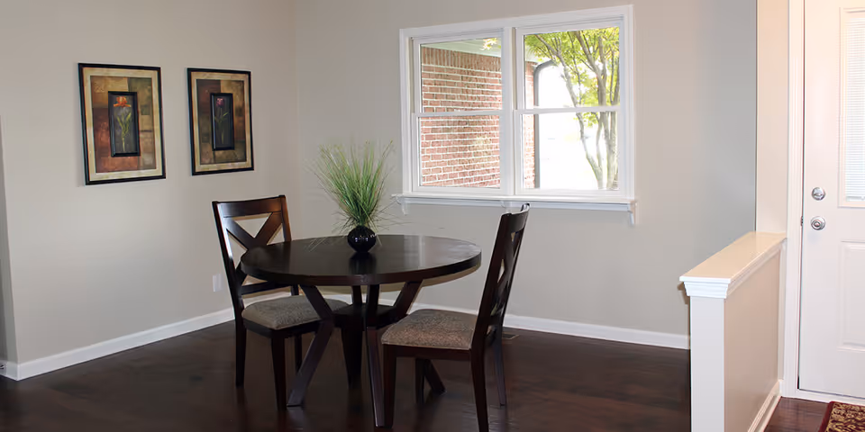 A small dining area with a round dark wooden table and two matching chairs with cushioned seats. A small black vase with green decorative grass is placed on the table. Two framed floral artworks hang on the beige wall to the left. A window shows a view of a brick wall and a tree outside. A white door with a window is visible on the right side.