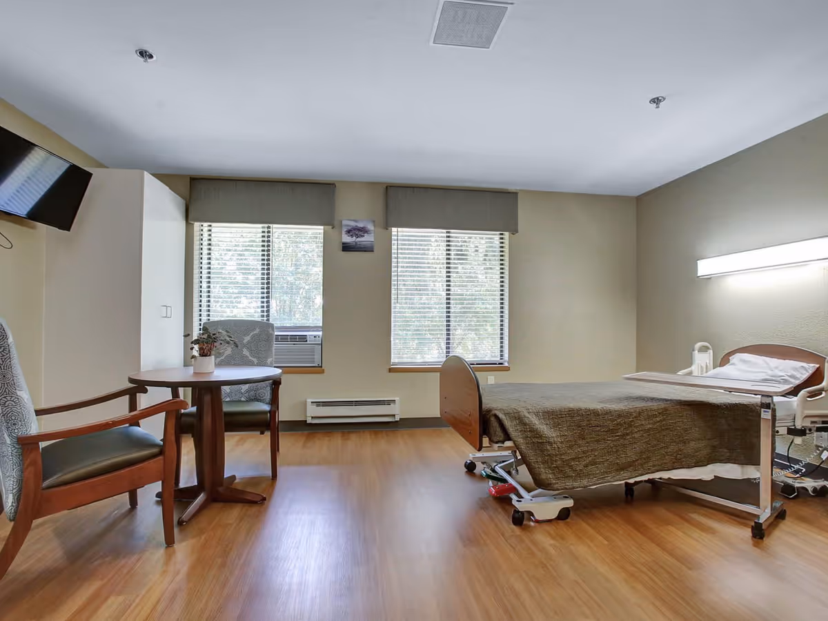A bright senior care room with a hospital-style bed, small table and chairs, a wall-mounted TV and two large windows.