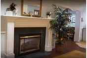 A cozy interior space featuring a white fireplace with a mirror above it, decorative items on the mantel, and a large potted plant to the right. The room has wooden flooring and an open doorway leading to another area with chairs and a table.