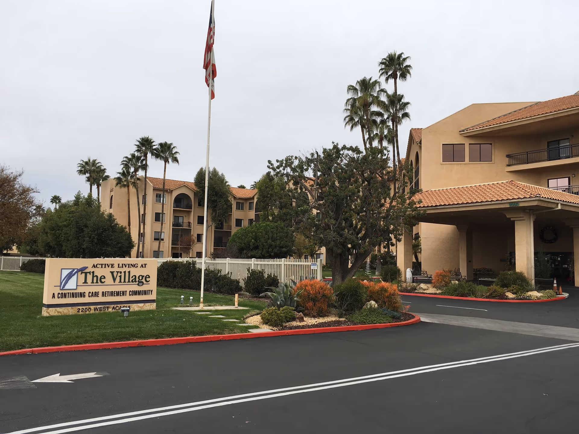 Exterior view of The Village HealthCare Center, a continuing care retirement community, showing a multi-story beige building with balconies, palm trees, a flagpole with an American flag, landscaped bushes, and a sign at the entrance with the facility name and address.