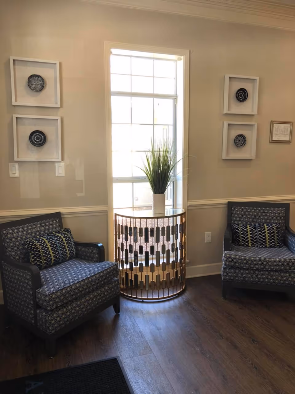 Two patterned armchairs flank a tall window with a round decorative side table holding a potted plant and framed wall art above.