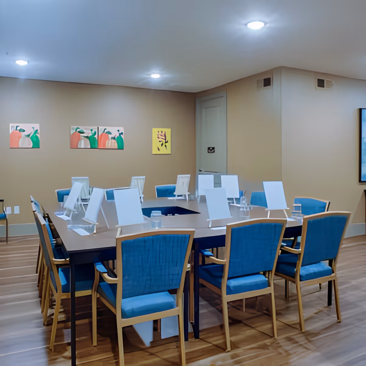 A conference or meeting room with a large rectangular table surrounded by blue cushioned chairs. The table has clear acrylic partitions and small water glasses placed on it. The room has beige walls with three colorful abstract paintings and recessed ceiling lights.