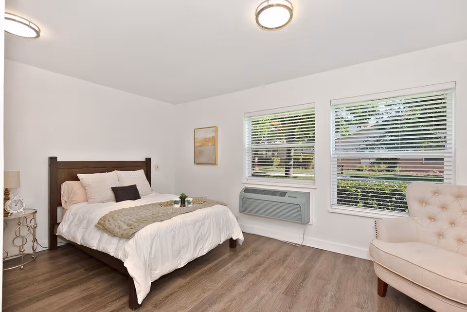 A bright and clean bedroom with a wooden bed frame, white bedding, and a beige throw blanket. There is a small round side table with a lamp and clock on the left side of the bed. Two large windows with white blinds let in natural light, and a beige upholstered armchair is positioned near the windows. The room has wood flooring and white walls with a framed picture hanging on one wall.