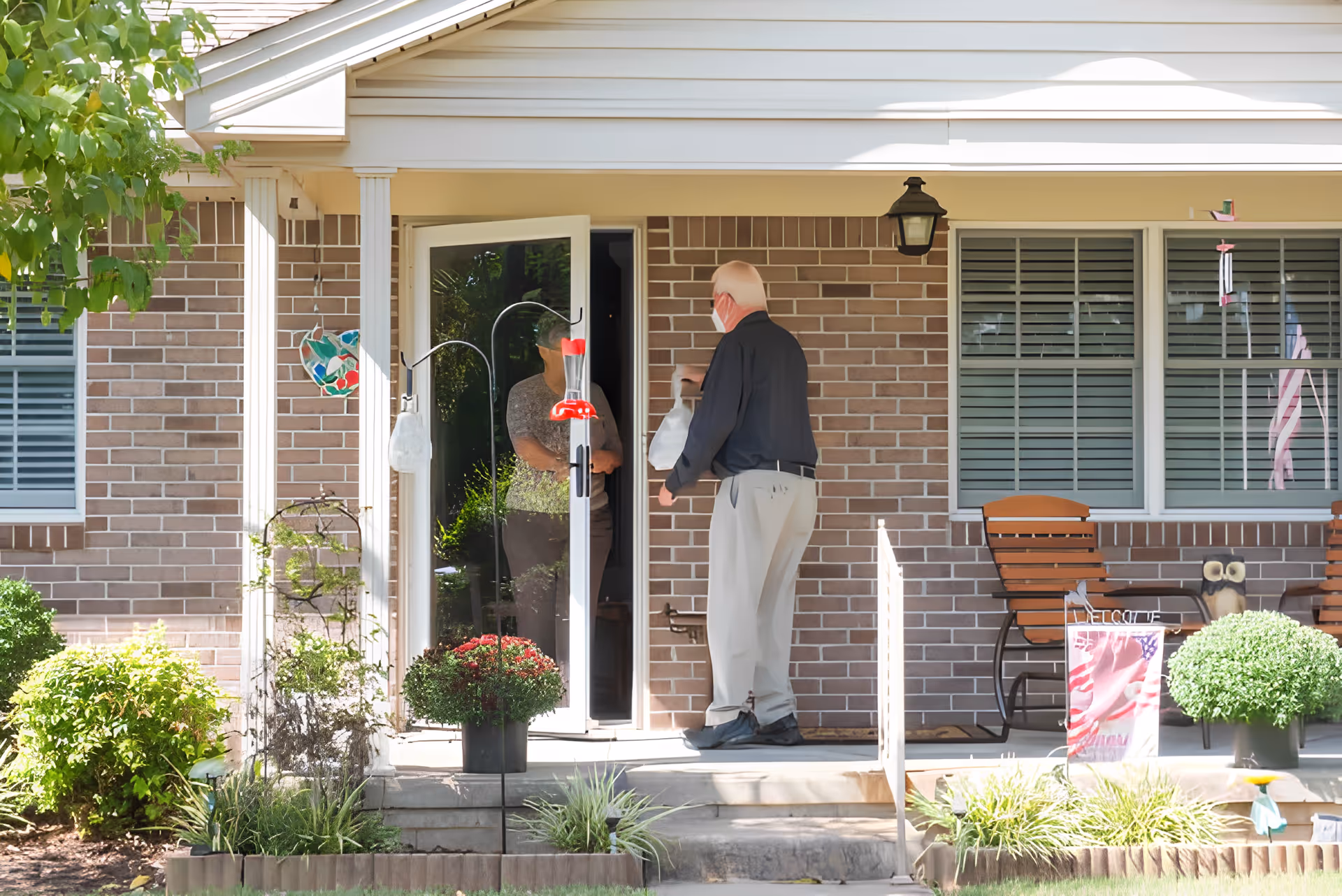 An elderly man wearing a mask is standing on the front porch of a brick house, handing a bag to a woman who is standing inside the open door. The porch has potted plants, two wooden chairs, and a small American flag decoration. There is greenery and a tree visible around the porch.