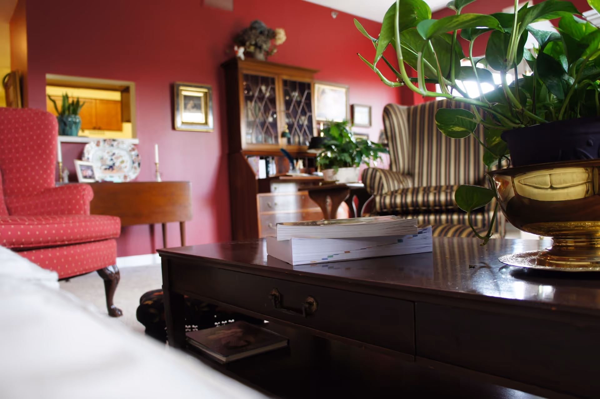 Cozy living room with a dark wooden coffee table in the foreground holding a stack of books and a potted plant in a golden pot. In the background, there is a red upholstered armchair, a wooden cabinet with glass doors, framed pictures on a red wall, and a striped armchair. The room has a warm and inviting atmosphere.