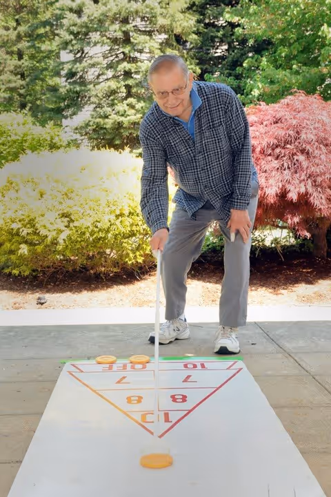 An elderly man wearing glasses, a blue checkered shirt, and gray pants is playing shuffleboard outdoors on a concrete surface. He is leaning forward holding a shuffleboard cue, aiming to slide a puck on the shuffleboard court. Behind him are green and red leafy bushes and trees.