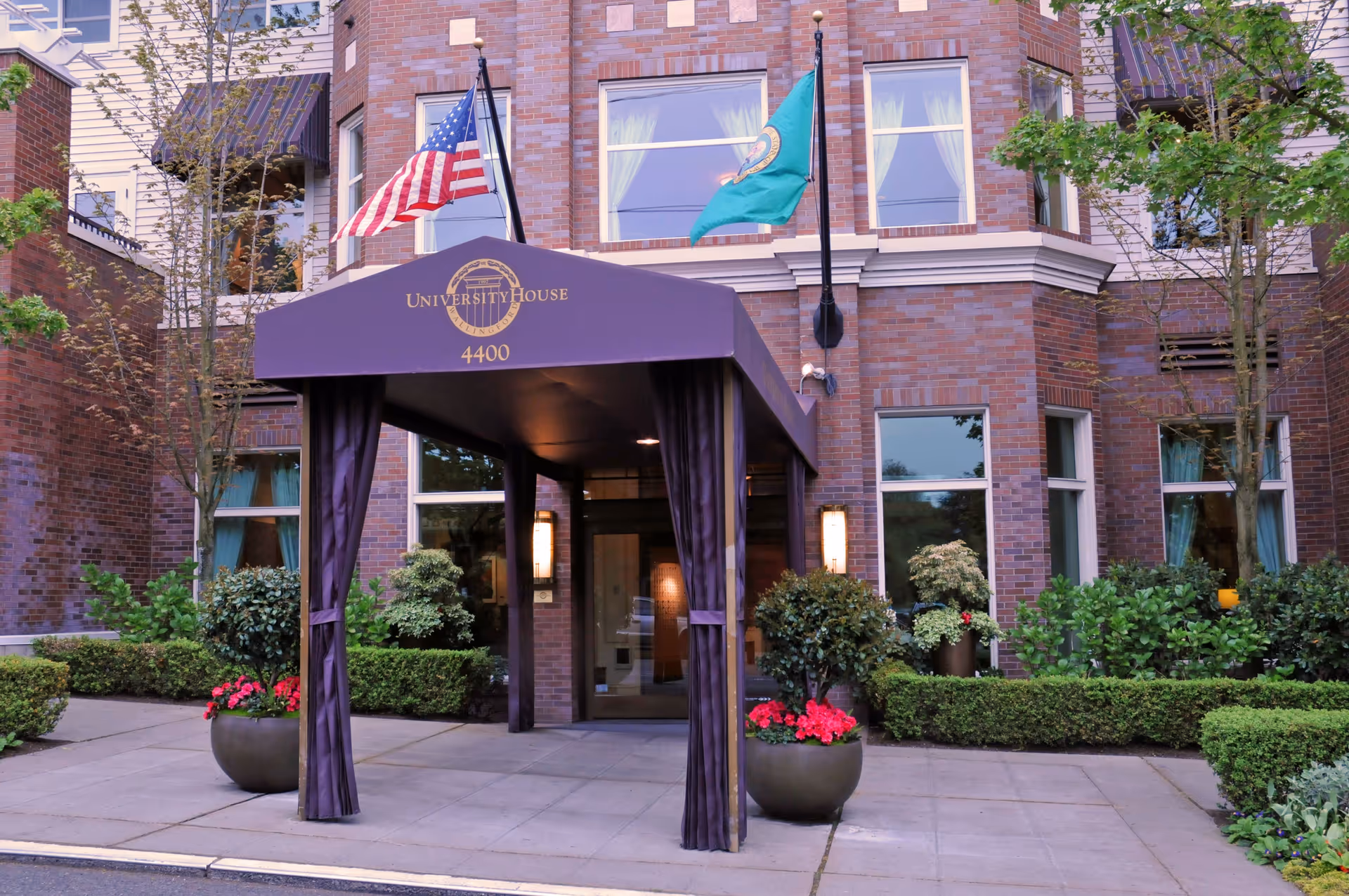 Entrance to University House Wallingford, a brick building with a covered walkway displaying the facility name and address 4400. Two flags, one American and one green flag, are mounted above the entrance. The area is landscaped with bushes, trees, and potted plants with red flowers.