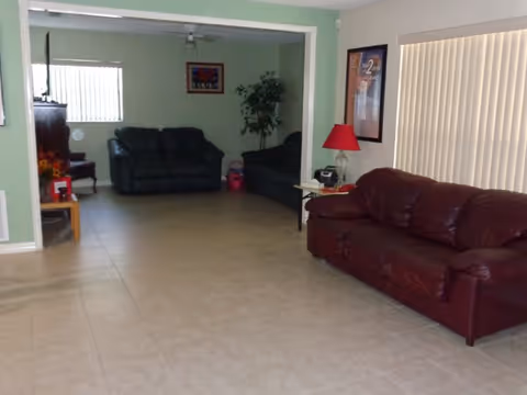 Interior view of a living room area with tiled floor, featuring a brown leather sofa on the right side, two dark-colored sofas in the background, a small side table with a red lamp, a framed picture on the wall, vertical blinds covering a window, and a potted plant in the corner.