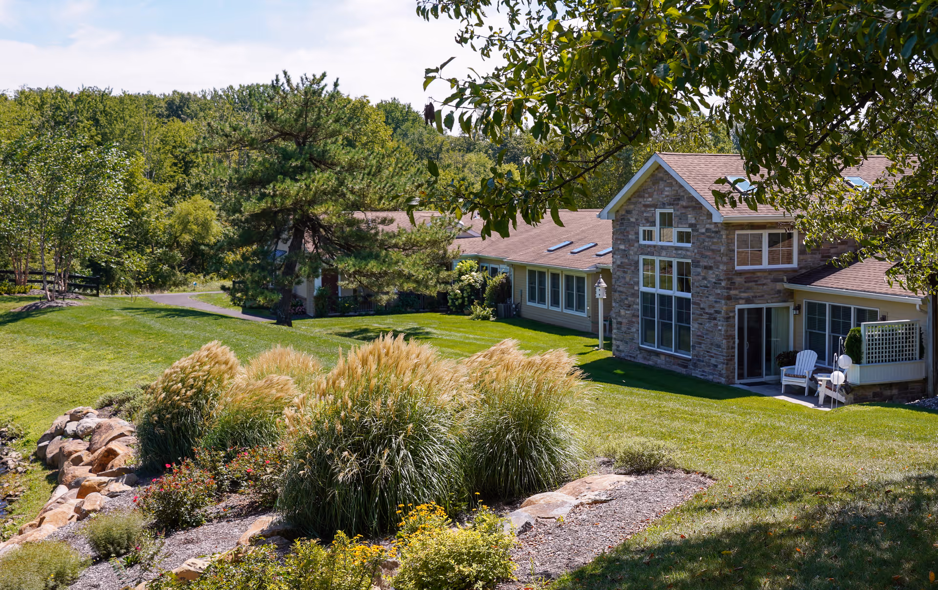 A scenic view of a senior living facility building with stone and siding exterior surrounded by lush green grass, ornamental grasses, flowering plants, and trees under a partly cloudy sky.