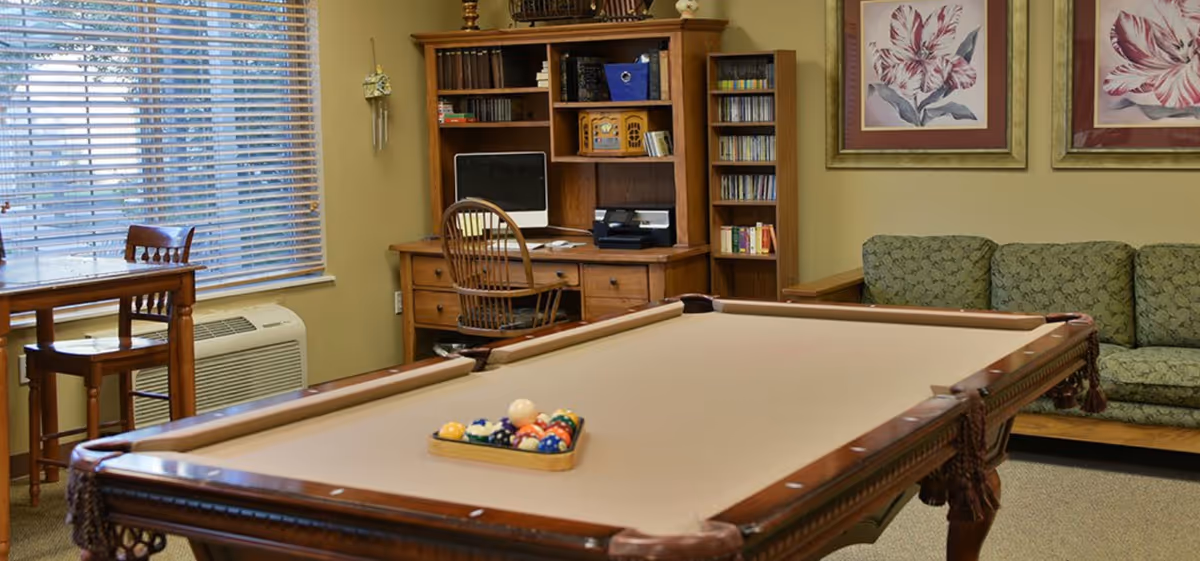 A cozy room featuring a beige pool table with a rack of billiard balls in the center. Behind the pool table is a wooden desk with a computer, printer, and shelves filled with books and CDs. To the right, there is a green patterned couch beneath two framed floral paintings. A wooden table with two chairs is positioned near a window with blinds, and an air conditioning unit is installed below the window.