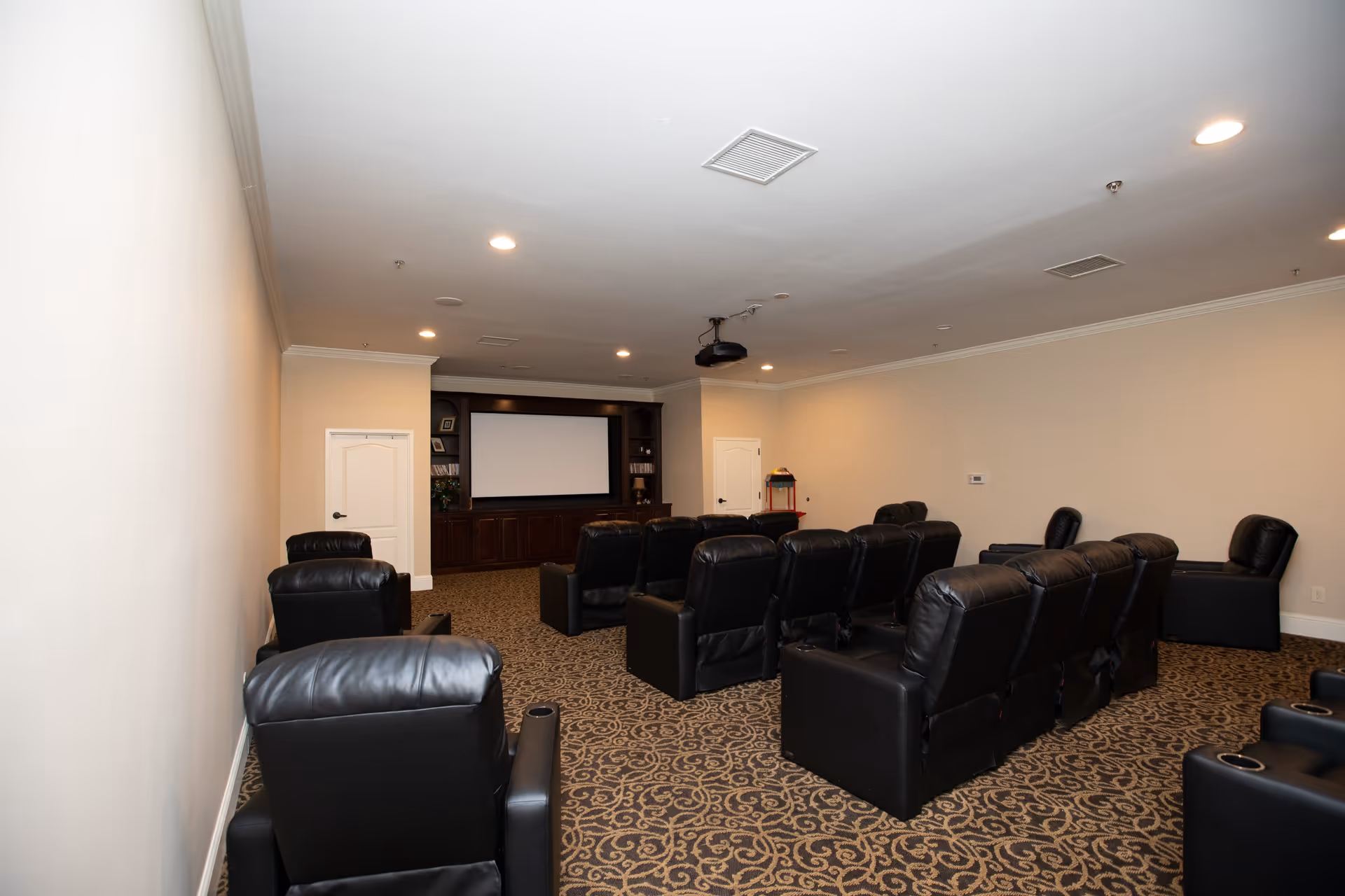 A small theater room with multiple rows of black leather recliner chairs facing a large projector screen mounted on a dark wooden entertainment unit. The room has beige walls, patterned carpet, recessed ceiling lights, and a popcorn machine in the back corner.