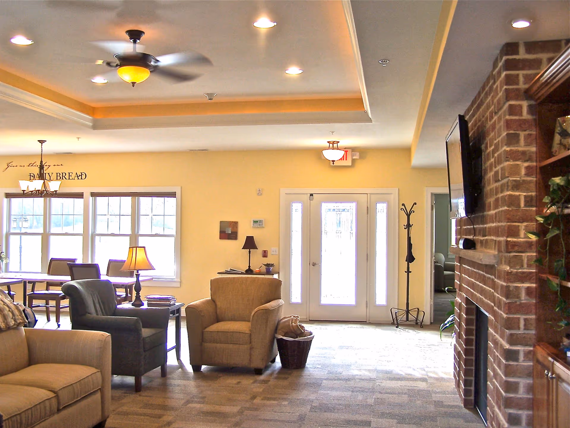 A cozy living room area in Maplewood Cottage featuring beige and dark armchairs, a beige sofa, a brick fireplace with a mounted TV, a coat rack, and a dining area with a table and chairs near large windows. The walls are painted light yellow, and the ceiling has recessed lighting and a ceiling fan.
