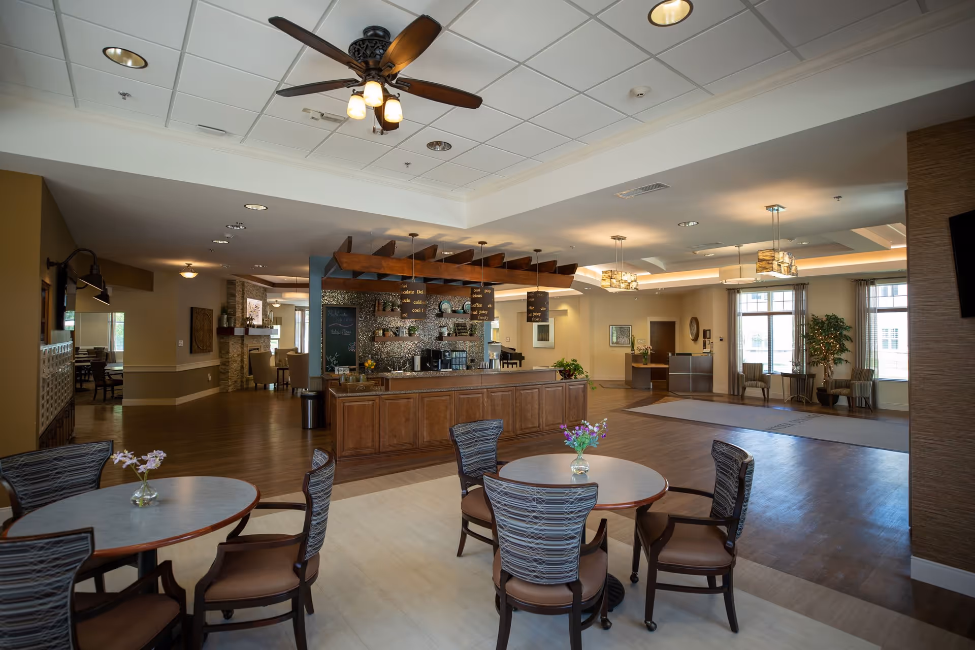 A spacious and well-lit common area in a senior living facility featuring round tables with chairs, a ceiling fan with lights, a wooden counter with hanging wooden beams and pendant lights, and large windows allowing natural light. The area has a warm and inviting atmosphere with a mix of wood and neutral tones, plants, and comfortable seating.