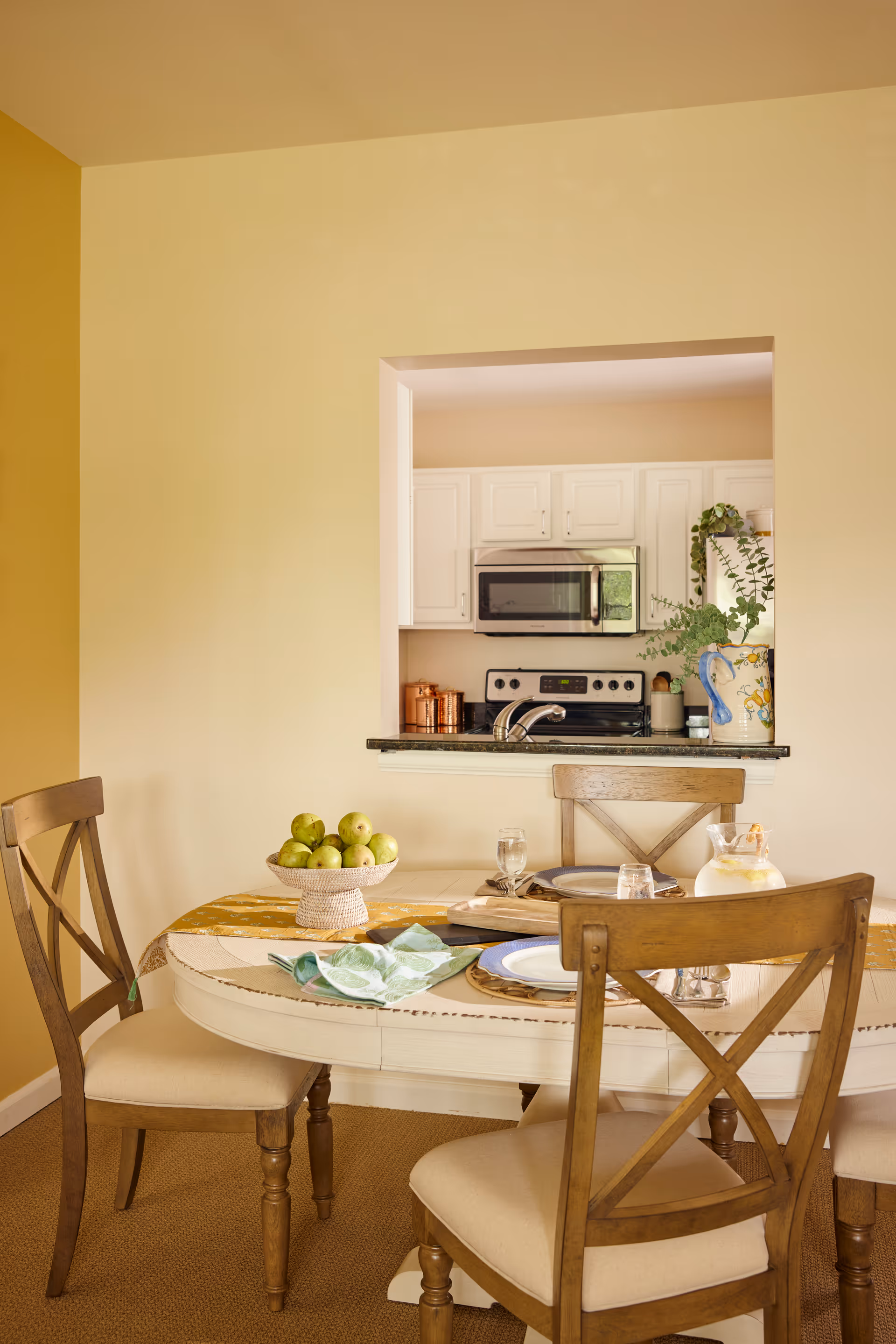 A dining area with a round table set for a meal, featuring wooden chairs with beige cushions. On the table, there is a bowl of green apples, plates, glasses, and a pitcher. In the background, a kitchen with white cabinets, a microwave, and a stove is visible through a pass-through window.