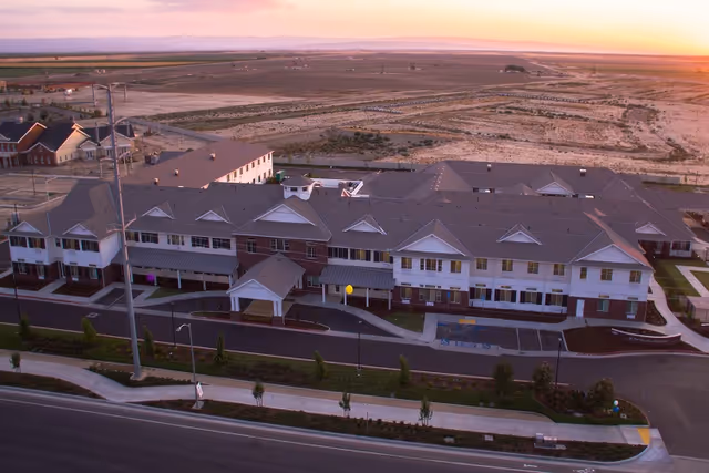 Aerial view of a large two-story senior living facility building named Ivy Park at Seven Oaks during sunset, surrounded by open land and a few other buildings in the distance. The building has a gray roof, white and brick exterior walls, and a covered entrance with a driveway in front.
