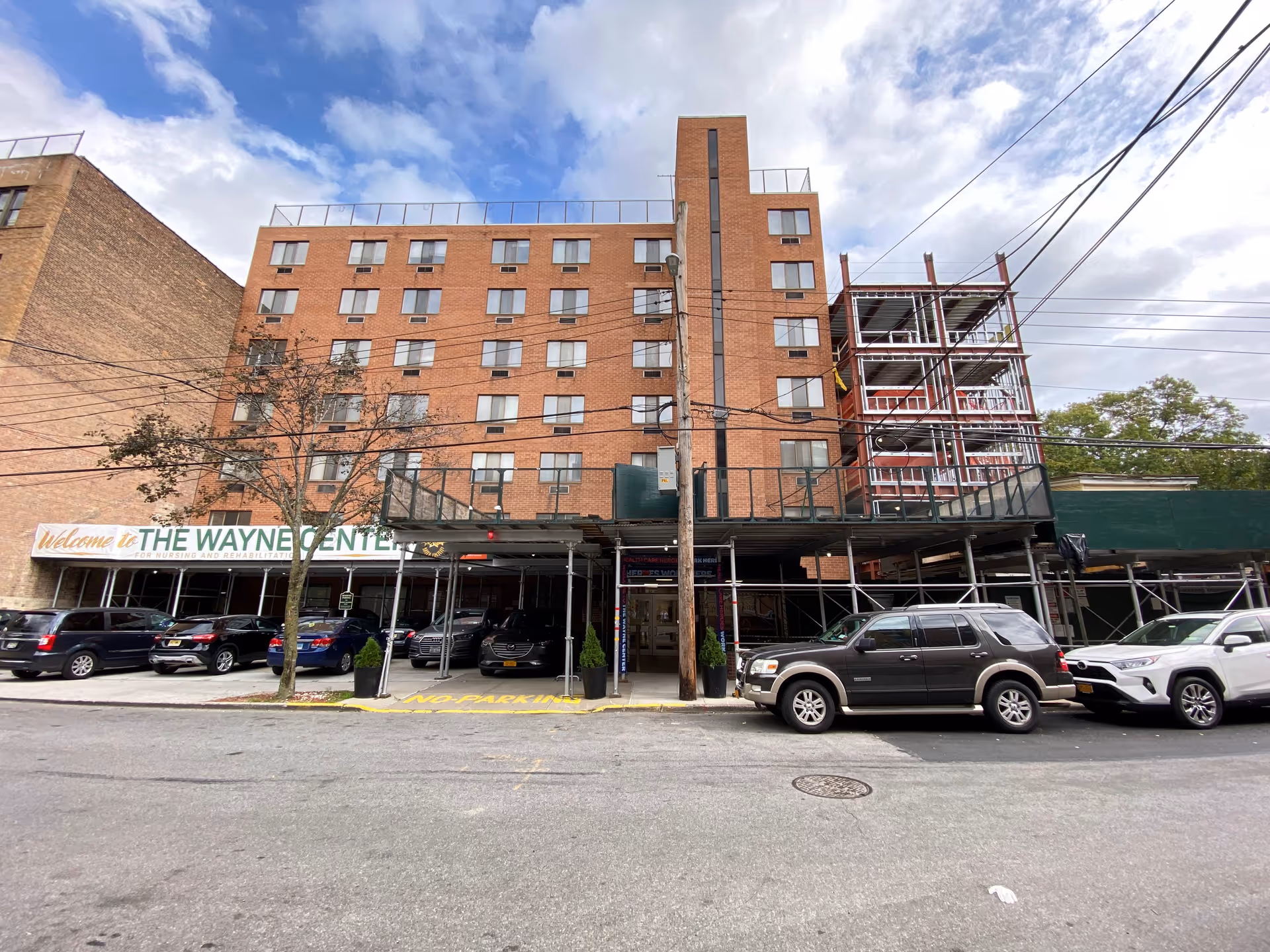 Exterior view of a multi-story brick building with several windows, scaffolding on the right side, and a covered entrance area. Several cars are parked in front of the building under the covered area and along the street. A sign above the entrance reads 'Welcome to THE WAYNE CENTER for Nursing and Rehabilitation.' The sky is partly cloudy.