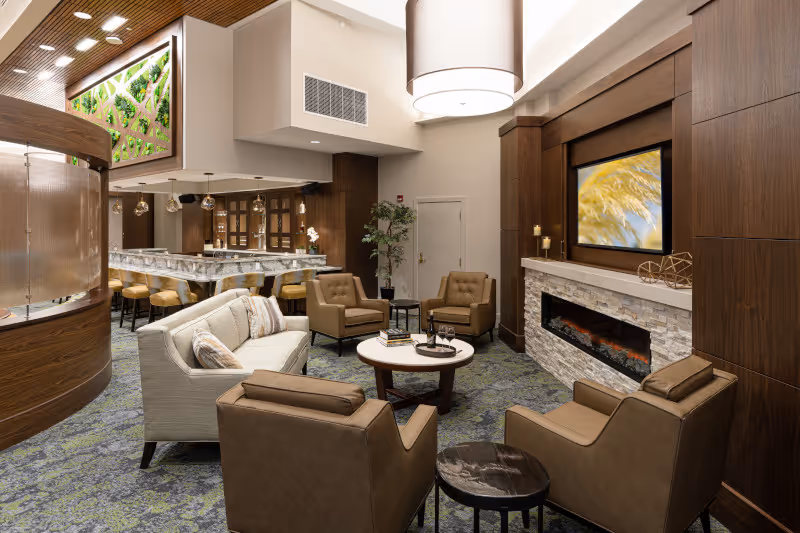 A modern and cozy living room area with a beige sofa, four brown armchairs arranged around a round coffee table, and a fireplace with a mounted TV above it. The room features wood paneling, a patterned carpet, and a bar area with high stools in the background. There is also a decorative green wall and pendant lighting above the bar.