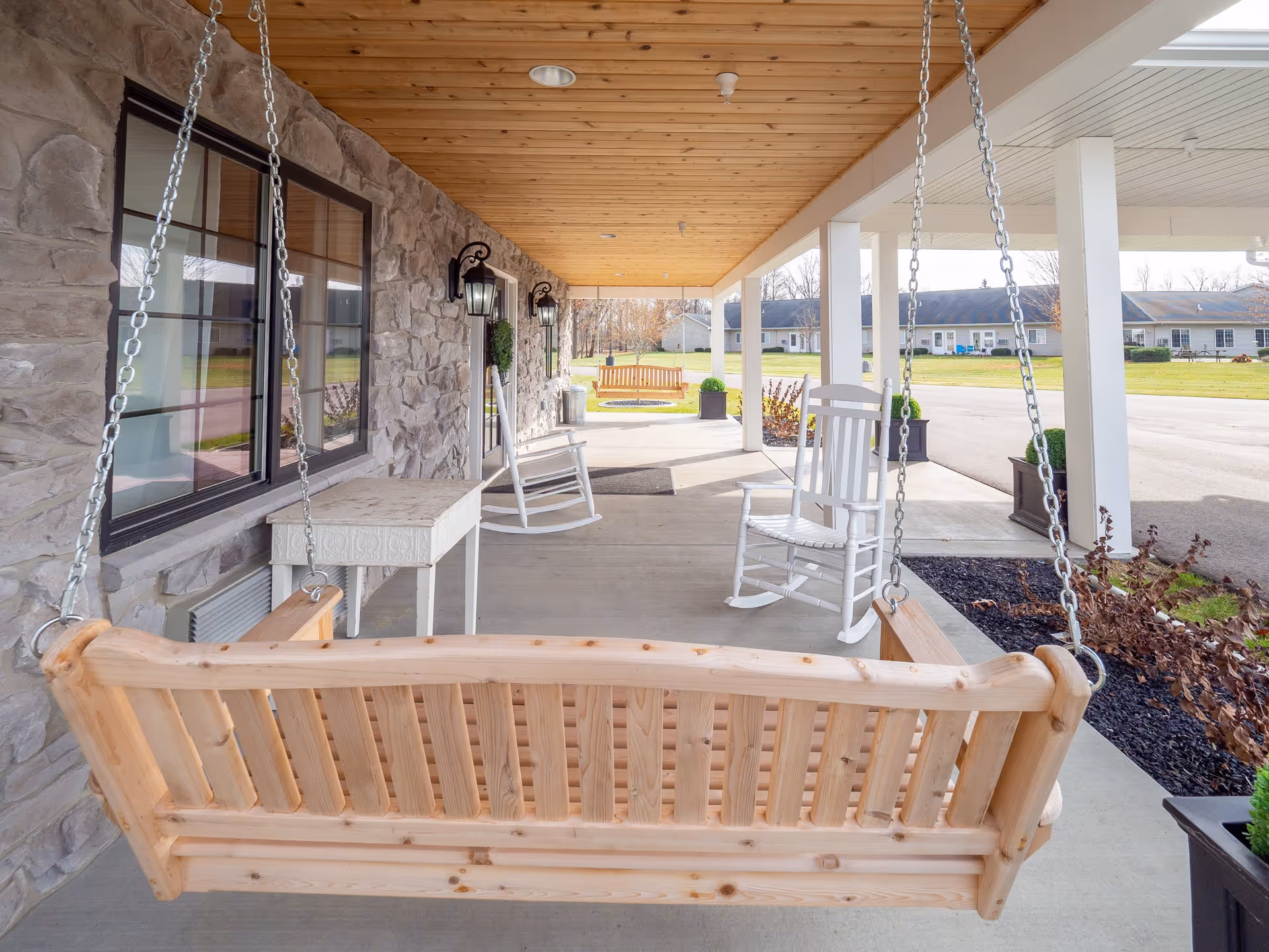 Covered front porch with a wooden hanging bench in the foreground, white rocking chairs, stone exterior wall, and columns overlooking the driveway.
