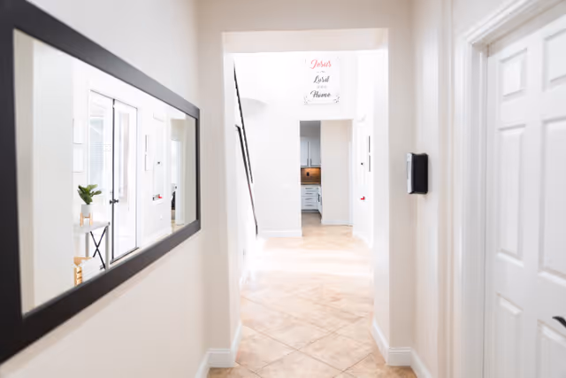 A bright hallway with beige tiled floor and white walls. On the left wall, there is a large rectangular mirror reflecting part of a room with a plant on a small table. At the end of the hallway, there is an open doorway leading to a kitchen area with white cabinets. Above the doorway, a sign reads 'Jesus is Lord of this Home'.