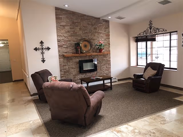A cozy living room area with two brown recliner chairs and one brown armchair arranged around a coffee table on a large rug. The room features a stone fireplace with a wooden mantel decorated with plants and a decorative plate. A large window with blinds lets in natural light, and a decorative metal cross hangs on the wall.