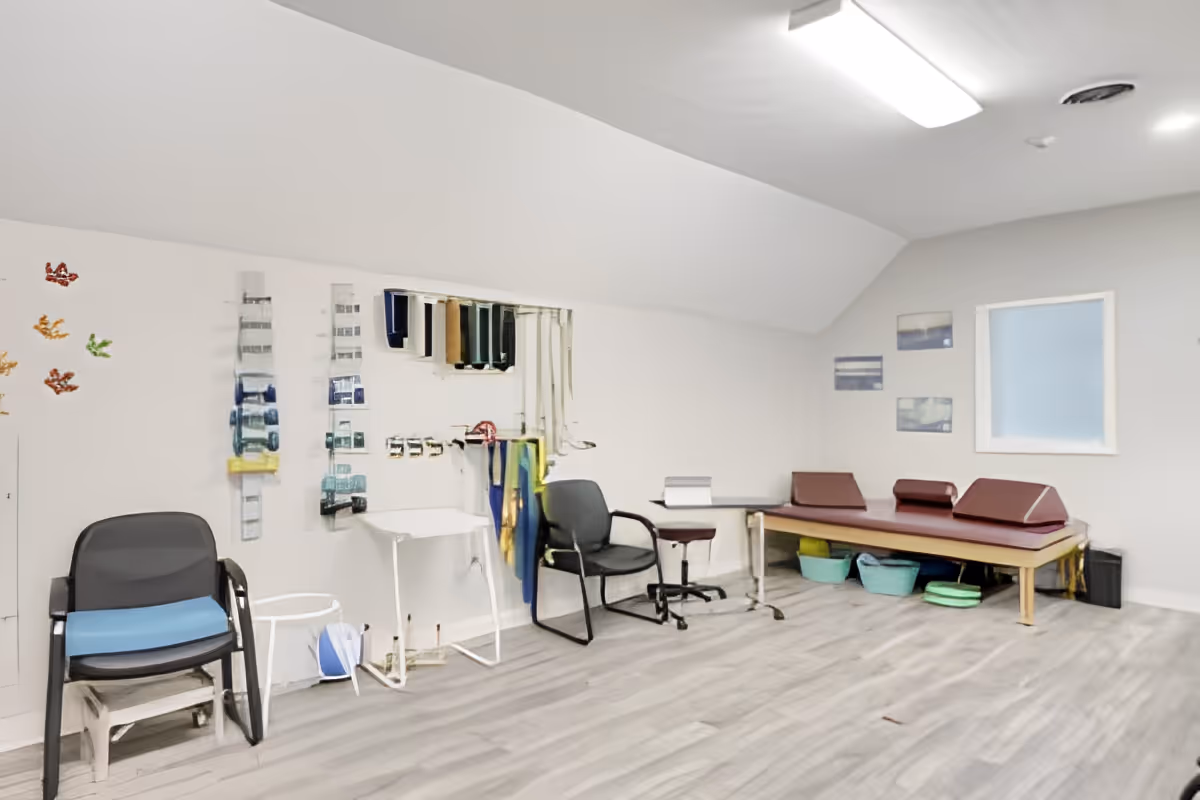 A therapy or rehabilitation room with exercise bands hanging on the wall, two chairs, a small table, and a padded therapy table with cushions. The room has light gray walls, wood-patterned flooring, and a window with frosted glass. There are a few framed pictures on the wall and some storage baskets under the therapy table.