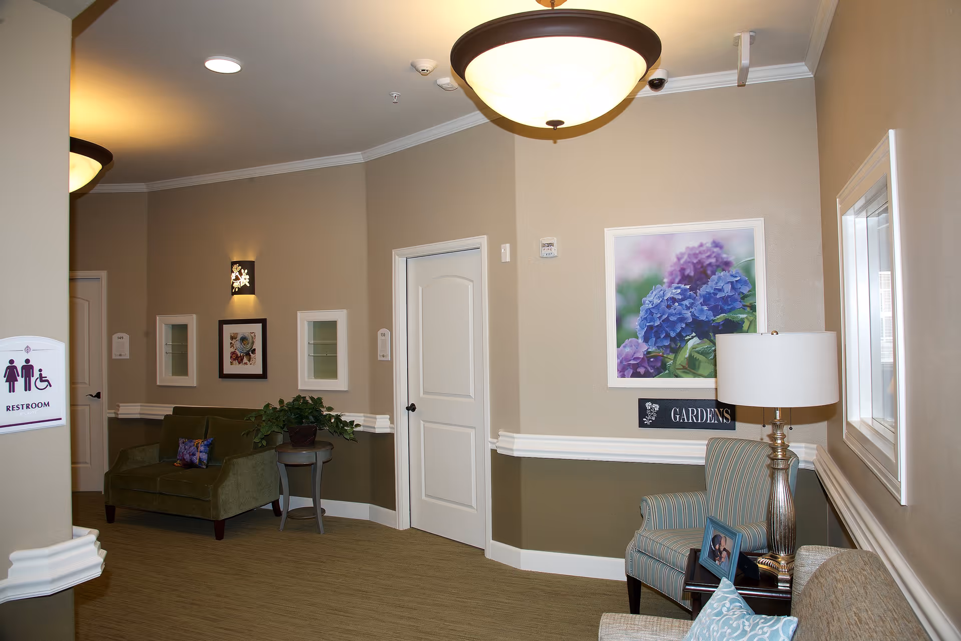 A cozy interior common area with beige and olive green walls, featuring a green loveseat with a decorative pillow, a small round table with a plant, a striped armchair next to a lamp and a framed photo, and a beige couch with a blue patterned pillow. There are framed pictures on the walls, including one of purple and blue flowers, and a sign indicating restrooms with accessibility symbols.