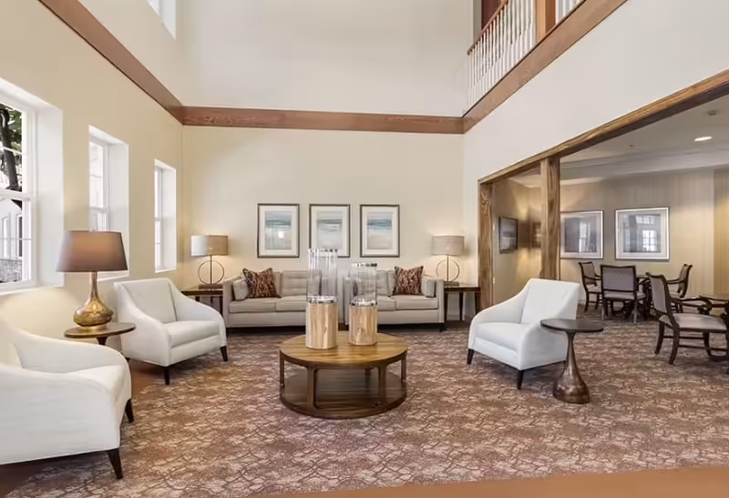 A spacious and well-lit living room area with a high ceiling and wooden railing above. The room features a beige sofa with decorative pillows, two white armchairs, a round wooden coffee table with decorative items, and two side tables with lamps. In the background, there is an adjoining room with a dining table and chairs, and framed artwork on the walls.