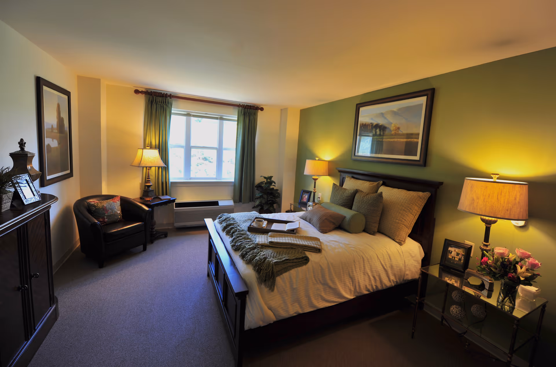 A cozy bedroom with a large bed made with white linens, multiple pillows, and a green throw blanket. There are two bedside tables with lamps, a framed picture above the bed, a window with green curtains, a black armchair, and a wooden cabinet. The room has warm lighting and a green accent wall behind the bed.