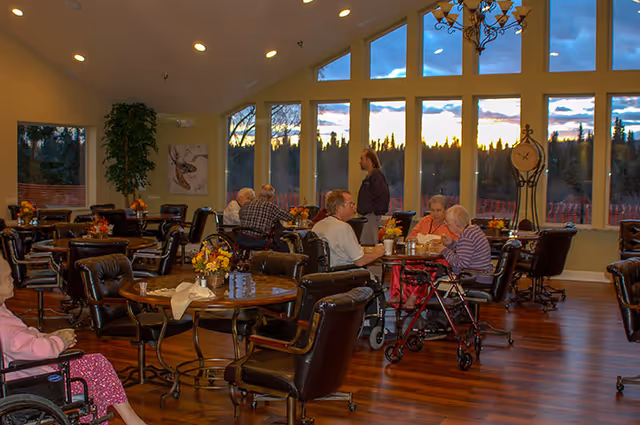 A spacious dining room with large windows showing a sunset outside. Several elderly people are seated at round tables with dark leather chairs, some using walkers or wheelchairs. The room has wooden floors, floral centerpieces on the tables, and a tall clock near the windows.