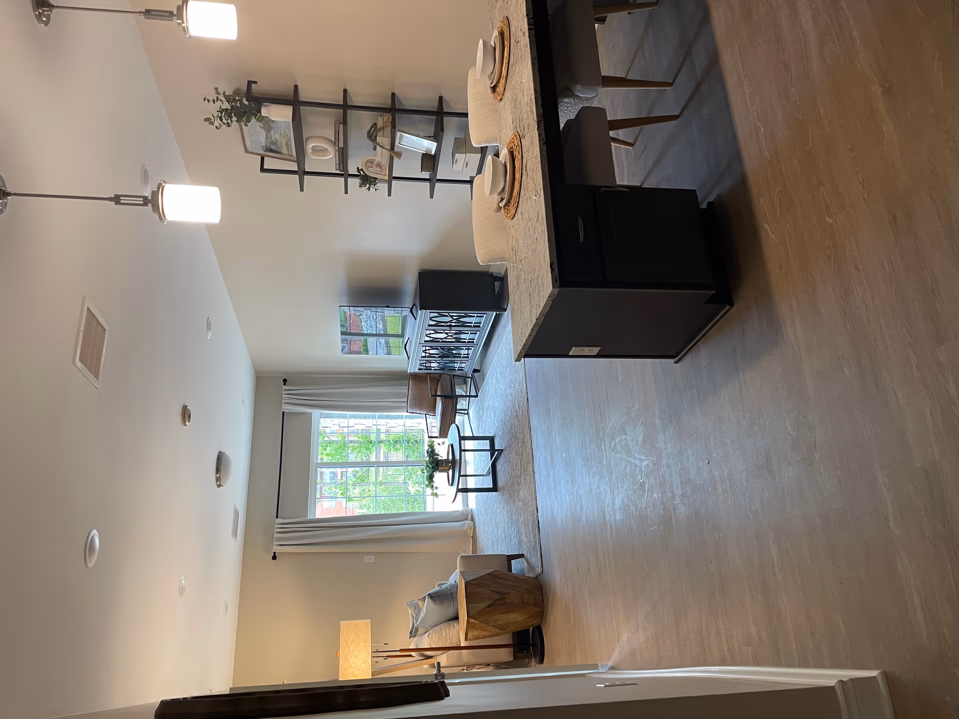 A bright and modern living room area with a large window covered by white curtains, a beige sofa with blue pillows, a wooden side table, a round coffee table with a plant, a leather chair, a TV on a black cabinet, and a black shelving unit with decorative items. In the foreground, there is a kitchen island with a granite countertop and two place settings with bowls and plates, along with two chairs. The floor is light wood, and the walls are painted beige.
