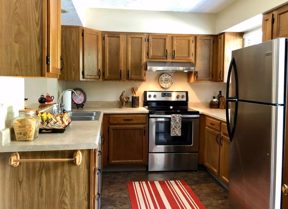 A kitchen with wooden cabinets, a stainless steel refrigerator and stove, a double sink, and a countertop with various items including a jar of pasta and a tray of snacks. A red and white striped rug is on the floor in front of the stove, and a clock with the word 'Coffee' hangs on the wall above the stove.