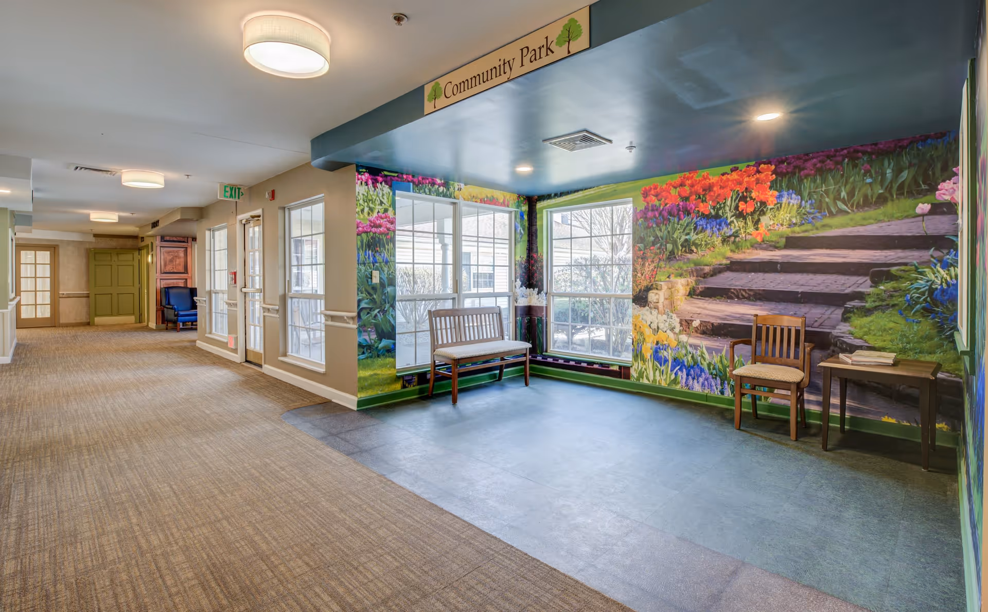 Interior hallway of a senior living facility with beige carpet and cream walls. At the end of the hallway is a seating area with a wooden bench and chair. The walls around the seating area feature a colorful mural of a garden with flowers and stone steps. Above the seating area is a sign that reads 'Community Park'. Large windows let in natural light.