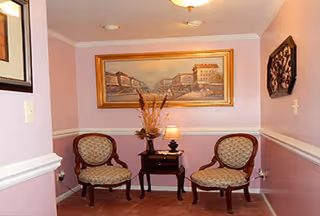 Small seating area with two upholstered chairs, a side table holding a lamp and floral arrangement, and framed artwork on the wall.