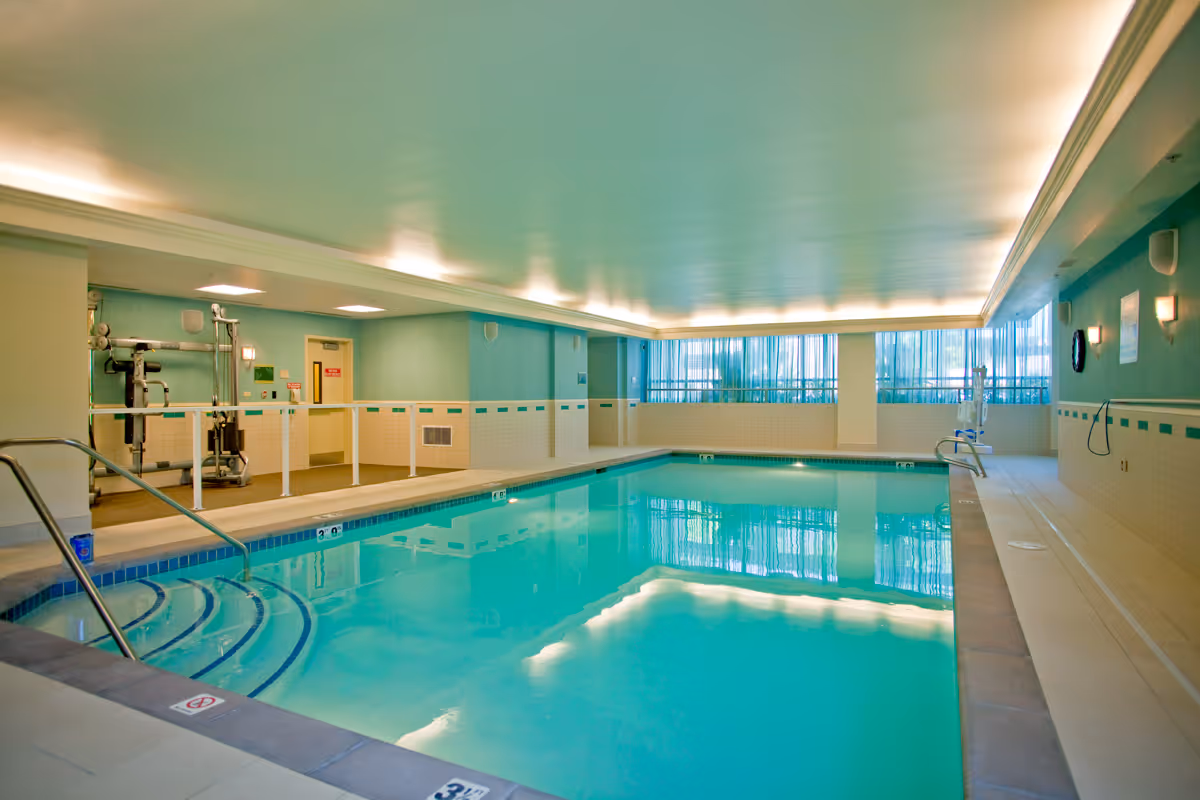 Indoor swimming pool with clear blue water, steps leading into the pool on the left side, and exercise equipment in the background. The room has teal walls, large windows with sheer curtains, and bright ceiling lights.