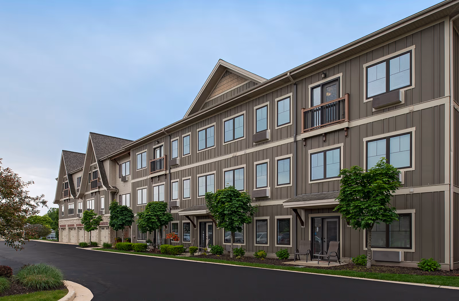 Front exterior of a three-story senior living apartment building with multiple windows, small balconies, and landscaped shrubs along a paved driveway.