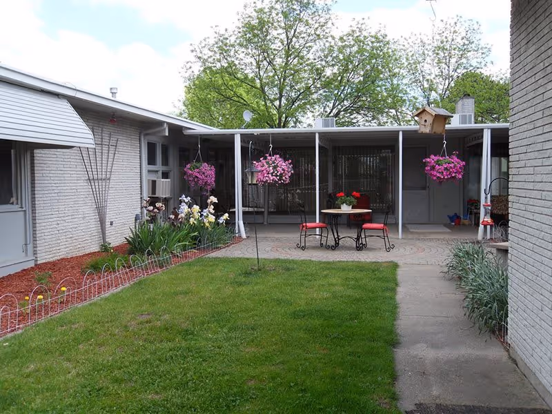 Enclosed courtyard with a grassy lawn, hanging pink flower baskets, a small patio table with red chairs, and single-story building wings.