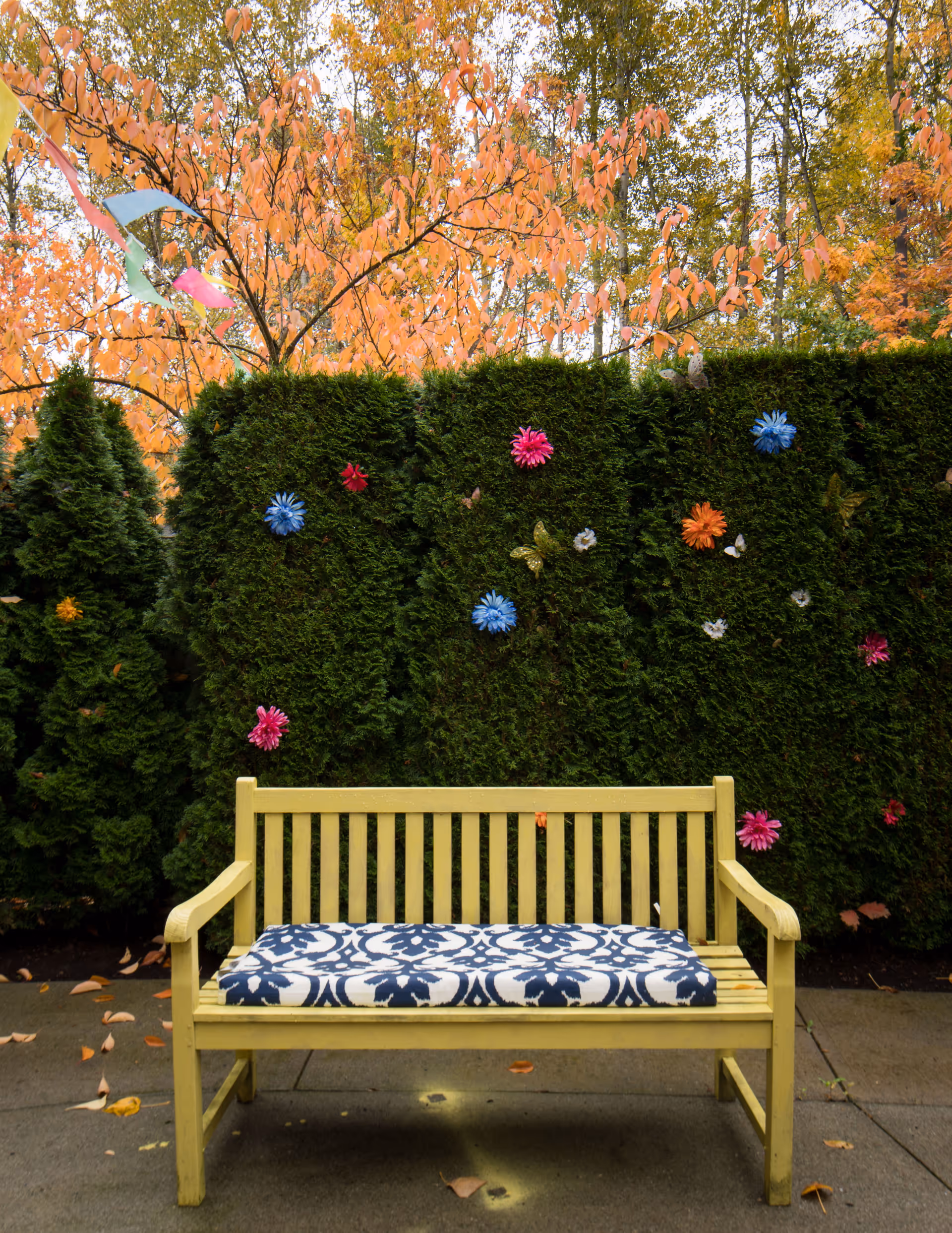 A yellow wooden bench with a blue and white patterned cushion is placed on a concrete surface in front of a tall green hedge decorated with colorful artificial flowers. Behind the hedge, trees with orange autumn leaves are visible.