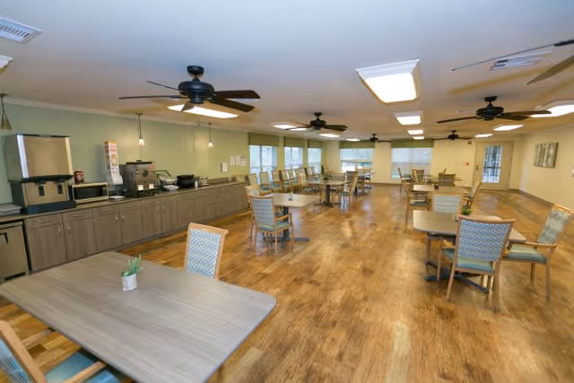 A spacious dining room with multiple tables and chairs arranged neatly on a wooden floor. The room features ceiling fans, large windows allowing natural light, and a long counter along one wall equipped with appliances including a microwave, coffee machine, and ice dispenser. The walls are painted light green and cream, and the room has a clean, welcoming atmosphere.