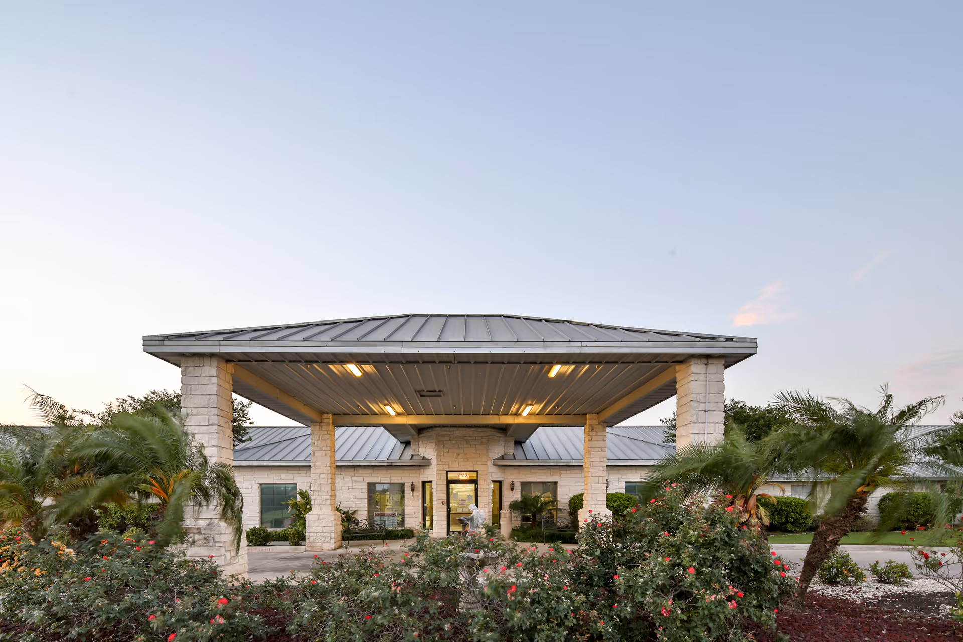 Entrance canopy of a single-story nursing and rehabilitation center with stone columns, a metal roof, landscaped beds and palm trees.