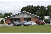 Single-story brick building with a peaked roof, several cars parked in front, and a grassy lawn.