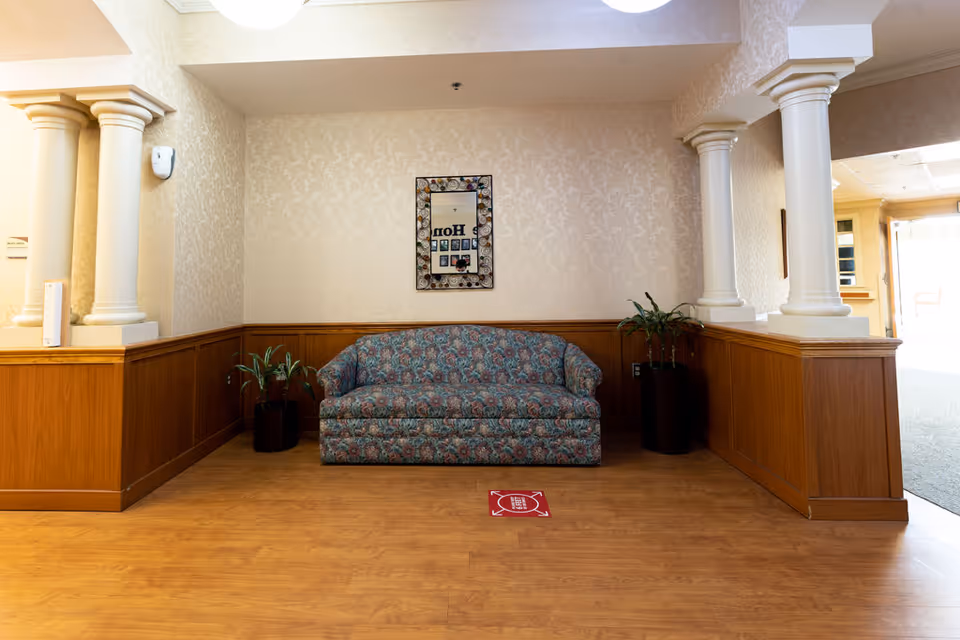 A small sitting area with a patterned blue and pink couch centered against a beige wallpapered wall with wooden wainscoting. Two potted plants flank the couch, and a decorative mirror hangs above it. The area is framed by white columns on either side, with wood flooring and a red floor marker in front of the couch.