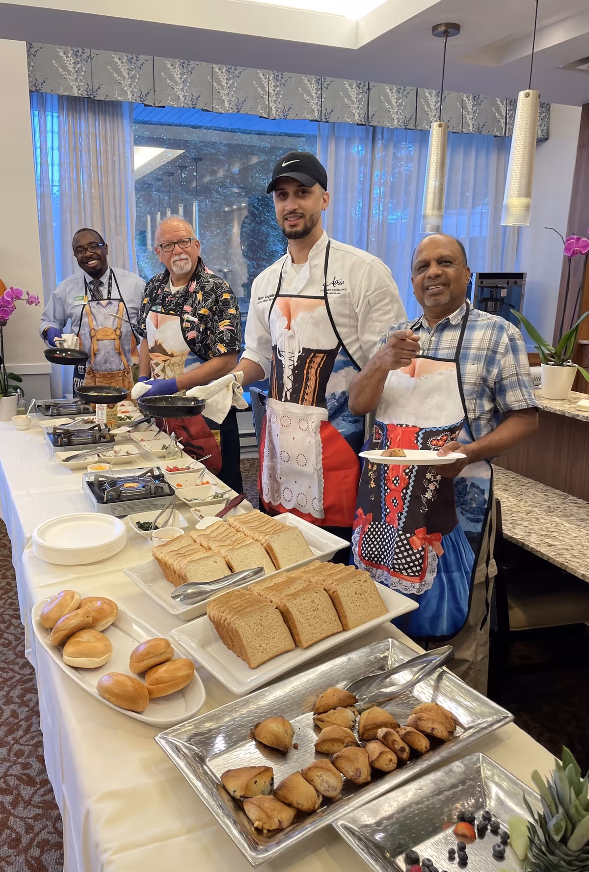 Four men standing behind a buffet table with various breads and pastries. They are wearing aprons and smiling at the camera in a well-lit room with large windows and hanging pendant lights.
