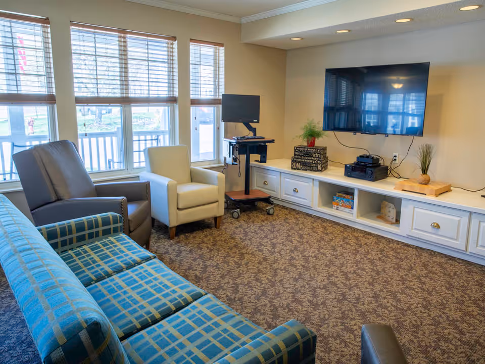 A cozy living room with a blue plaid sofa, a gray armchair, and a beige armchair arranged around a carpeted floor. A large flat-screen TV is mounted on the wall above a white built-in cabinet with drawers and open shelves. A small computer workstation on wheels is positioned near the window, which has wooden blinds partially open to let in natural light.