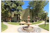 Outdoor courtyard with a multi-tier fountain, bench, pathways and apartment-style buildings surrounded by trees and lawn.
